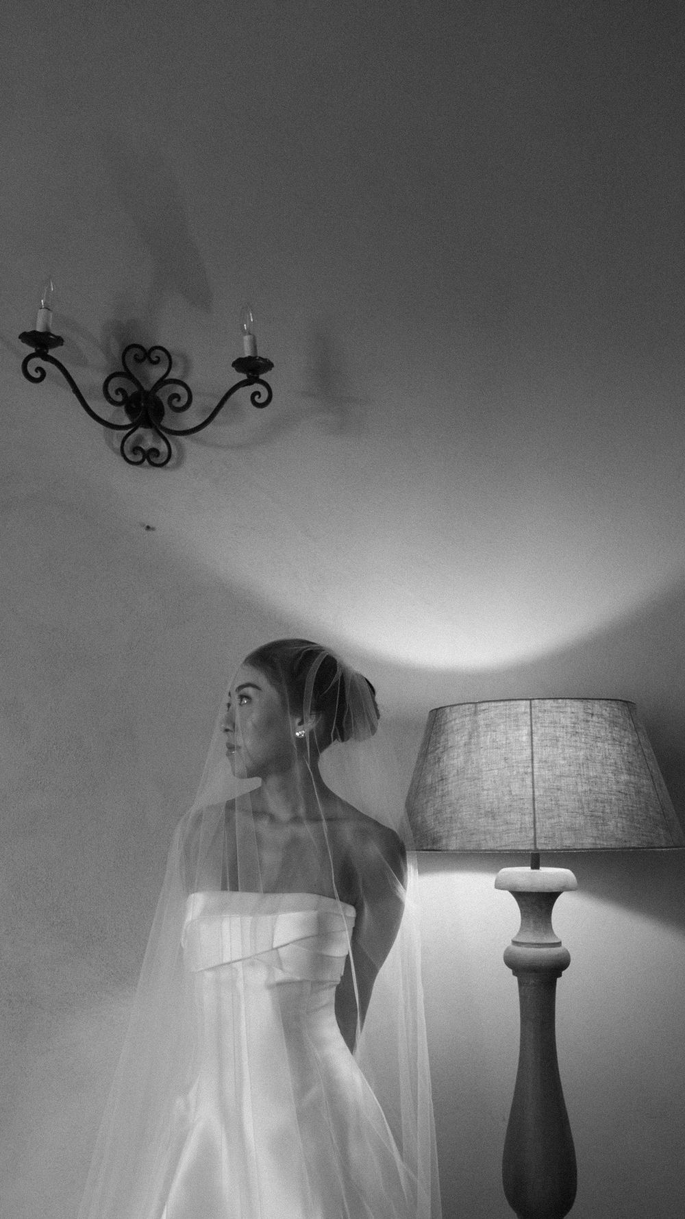 Black and white portrait of bride in strapless gown and veil standing beside lamp in dim room.