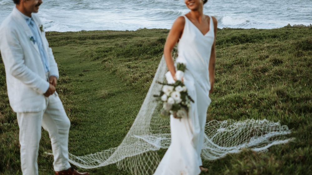 Bride and groom in white attire pose on coastal cliffside, bride holding bouquet with flowing net veil trailing behind.
