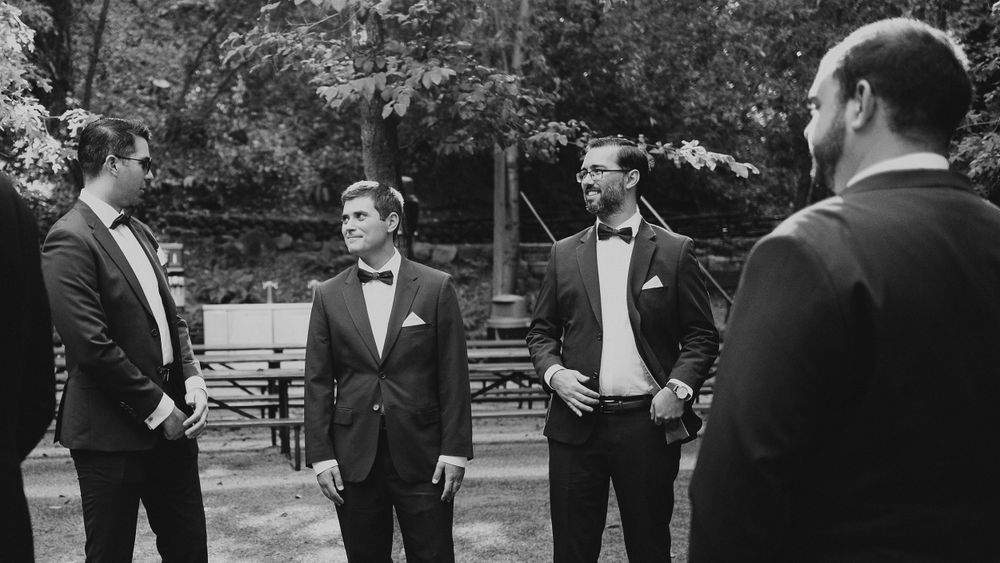 Black and white photo of groomsmen in tuxedos conversing outdoors at a wedding venue.