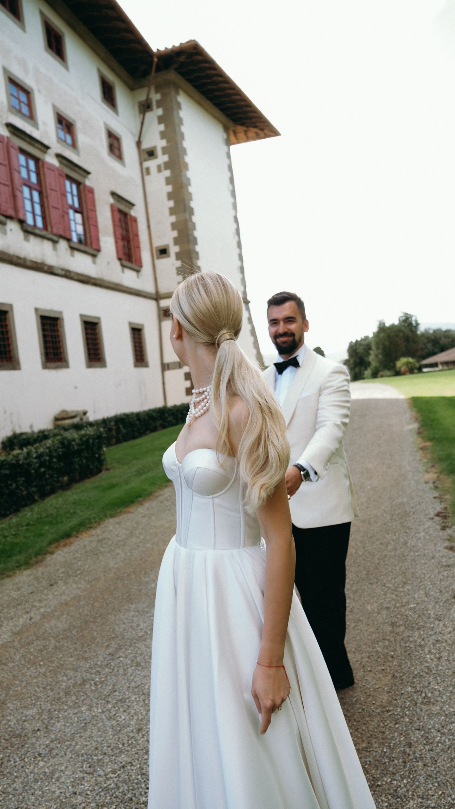 Bride in white strapless gown and groom in white tuxedo pose on gravel path outside elegant European villa.