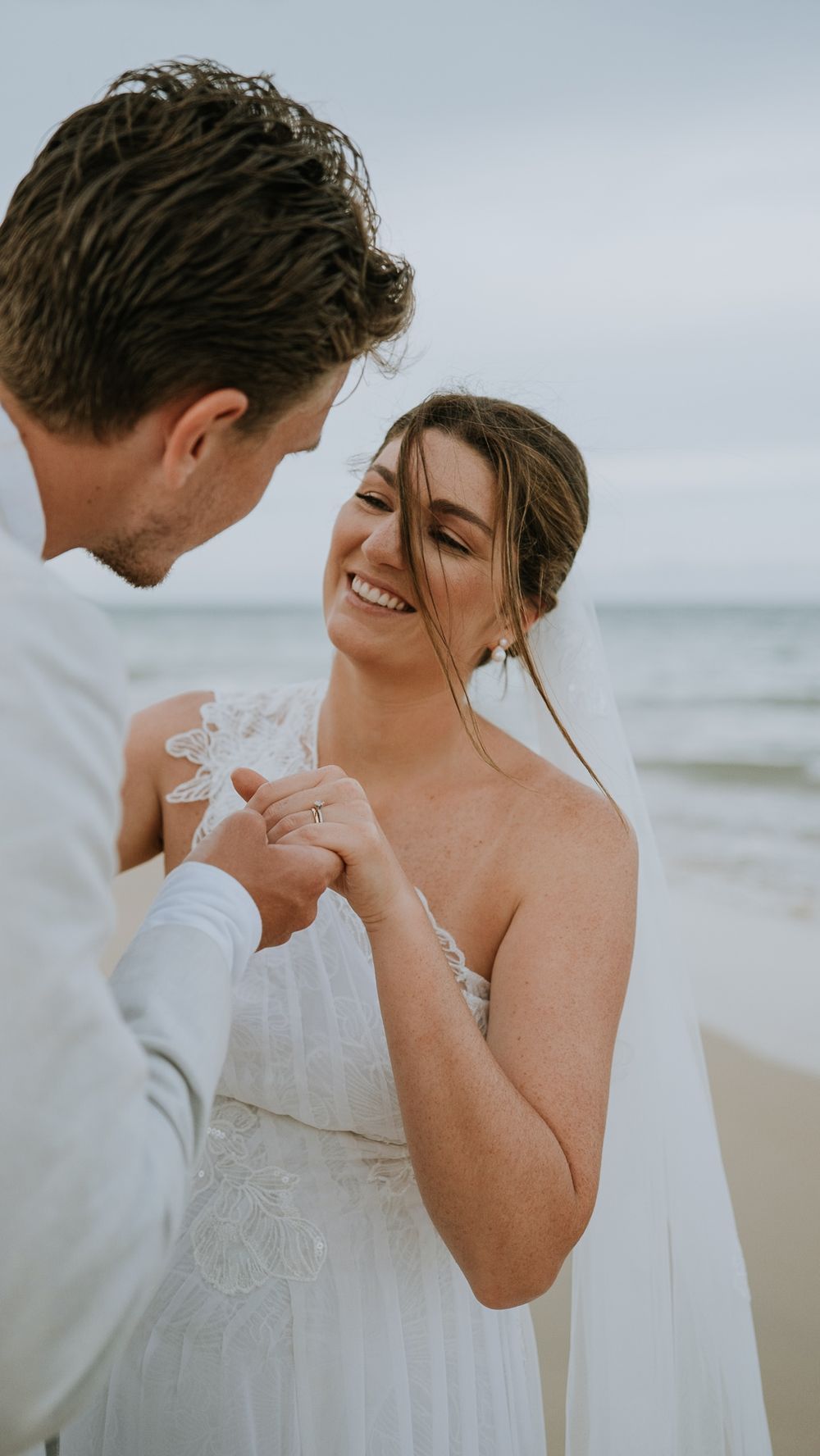 Smiling bride in white lace gown receives ring from groom on a beach wedding ceremony.