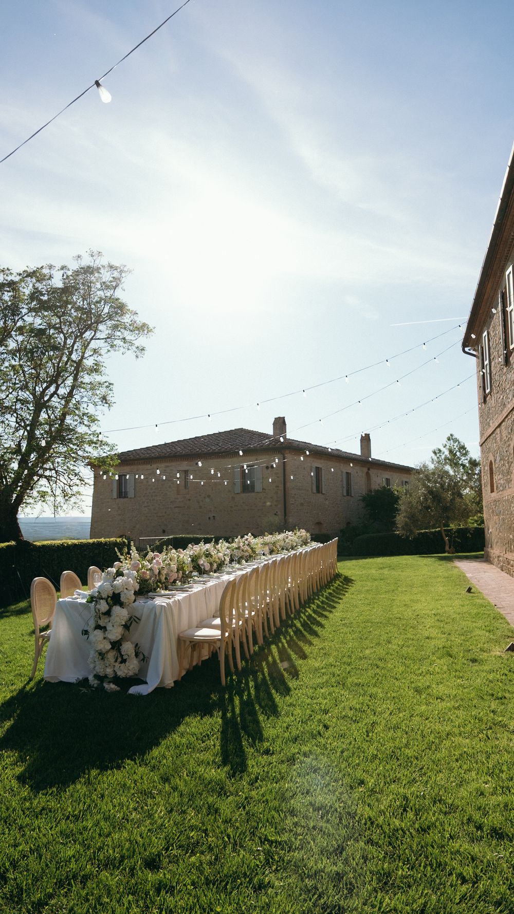 Elegant outdoor wedding reception with long floral table, string lights, and wooden chairs on lush green lawn.
