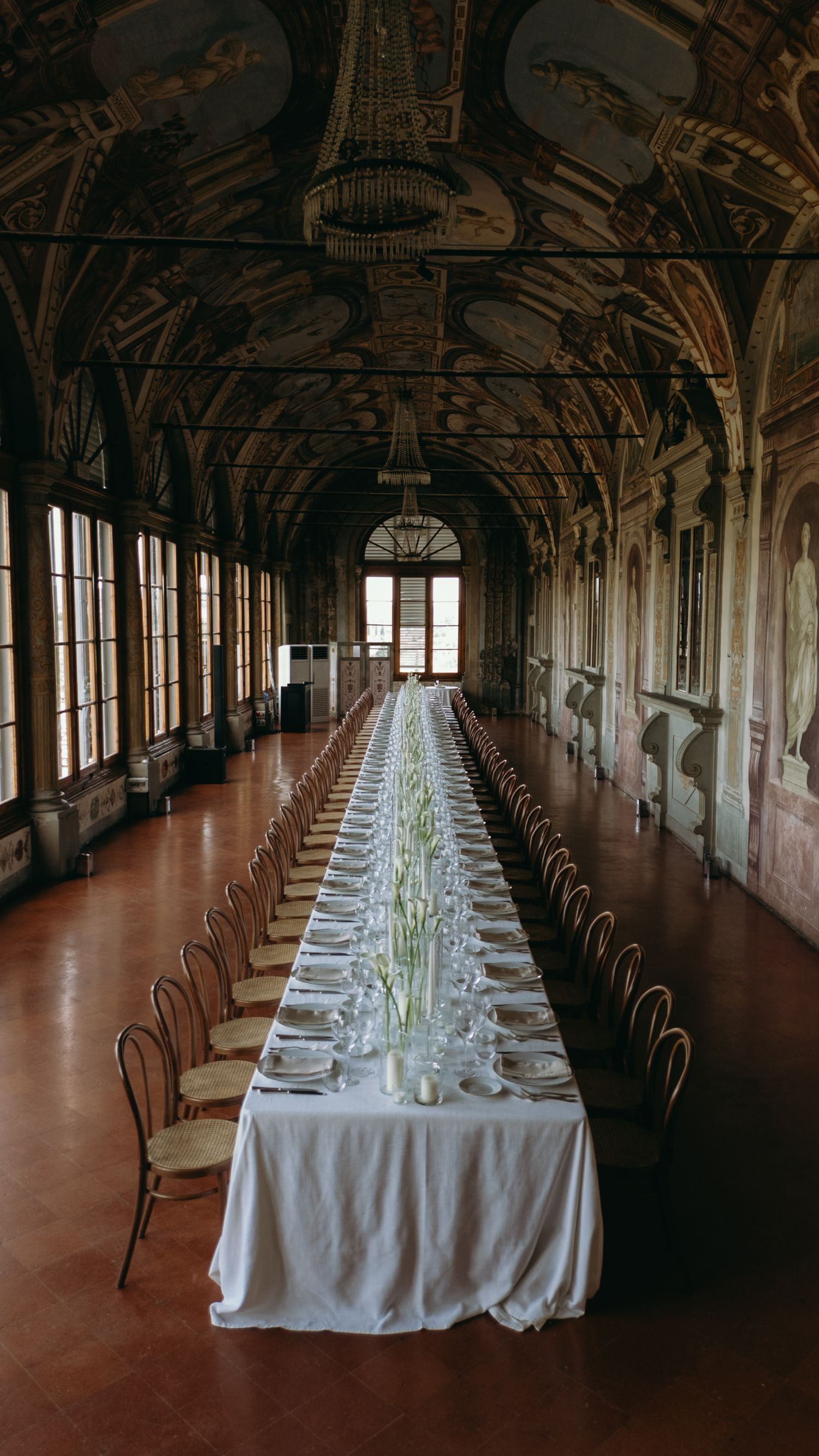 Elegant long banquet table with white linen set in an ornate Renaissance hall with frescoed ceilings and tall windows.