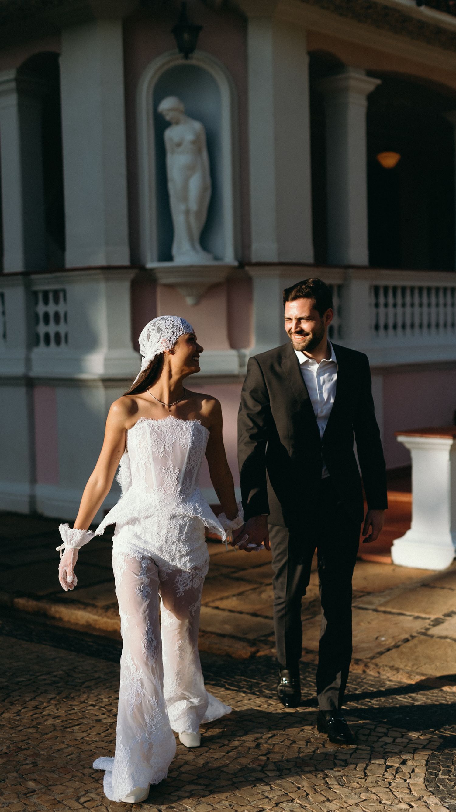 Bride in white lace pantsuit and groom in dark suit walking hand in hand at night wedding venue.