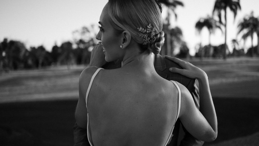 Black and white photo of bride in backless dress with floral hair accessory at outdoor wedding venue.