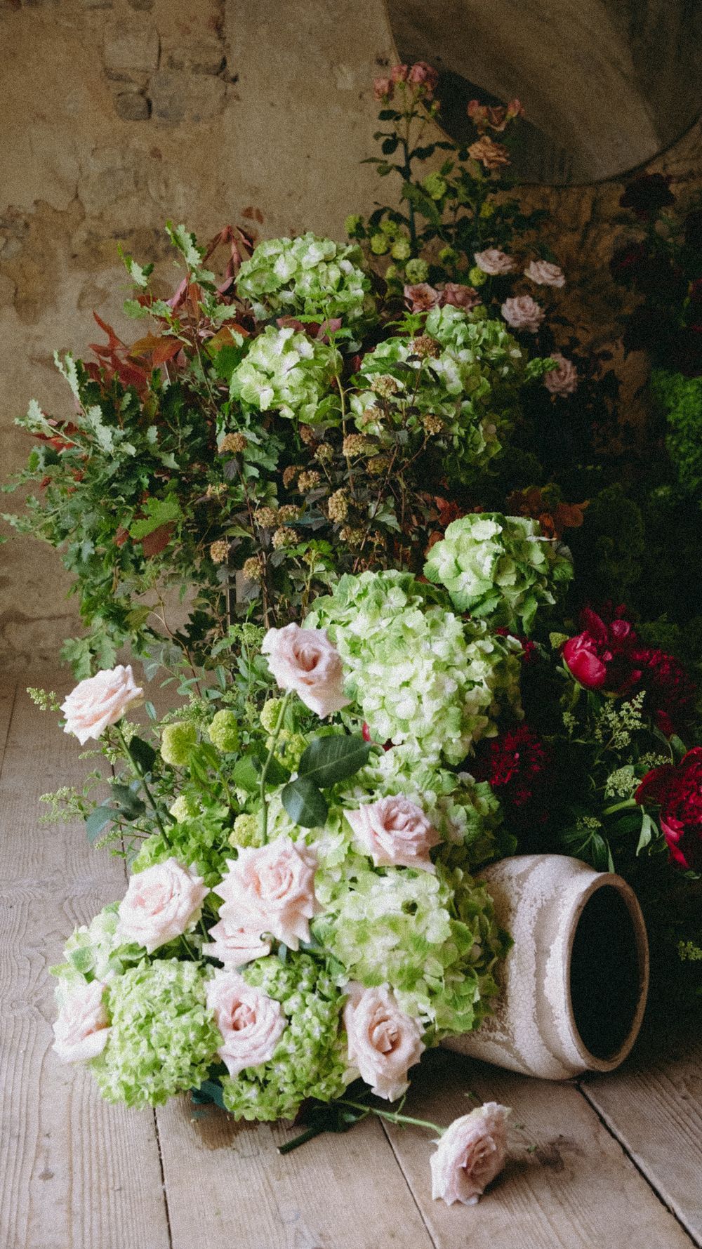 Lush floral arrangement of green hydrangeas, pink roses, and red flowers against a rustic stone wall.