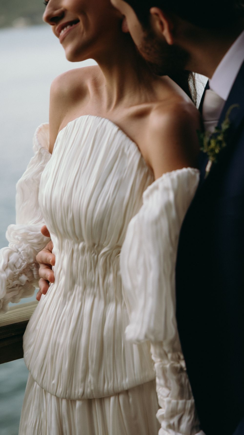 Bride in stunning off-shoulder pleated white wedding gown stands with groom near waterfront on wedding day.