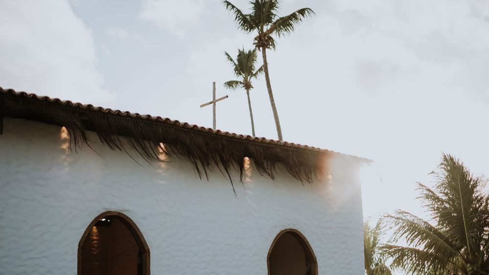 White chapel with thatched roof, cross, and palm trees against a bright sky in a tropical wedding venue.