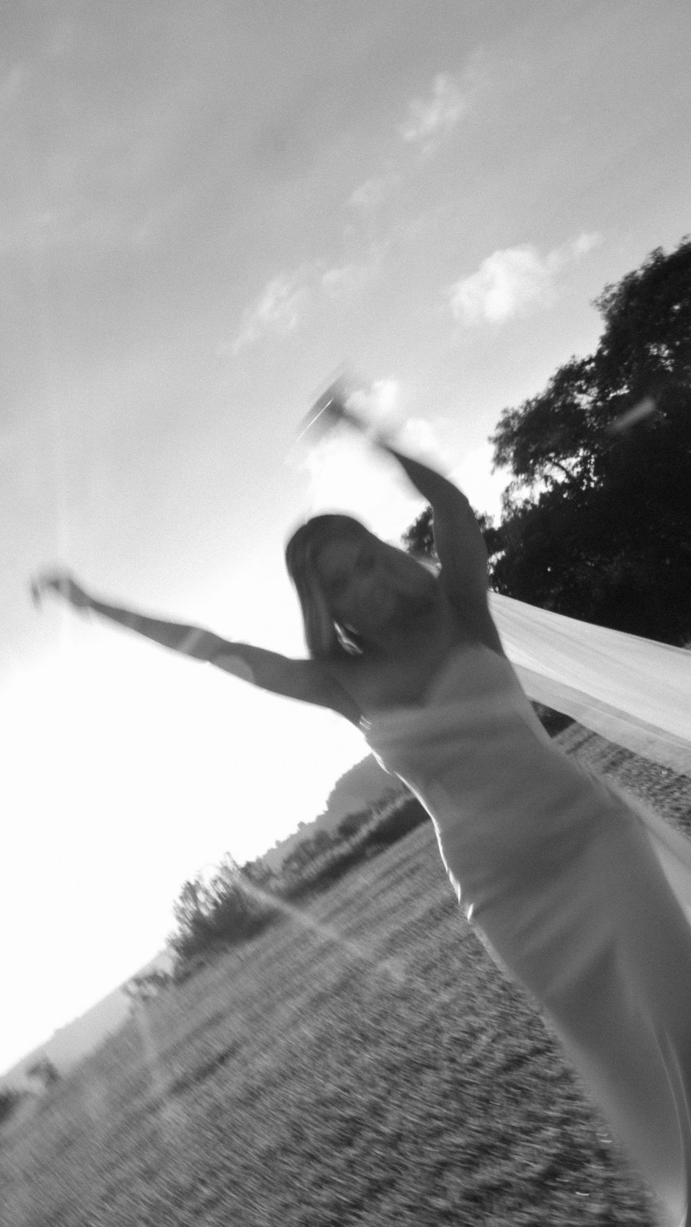 Bride in white gown twirls with arms outstretched in a sunlit black and white outdoor photo.