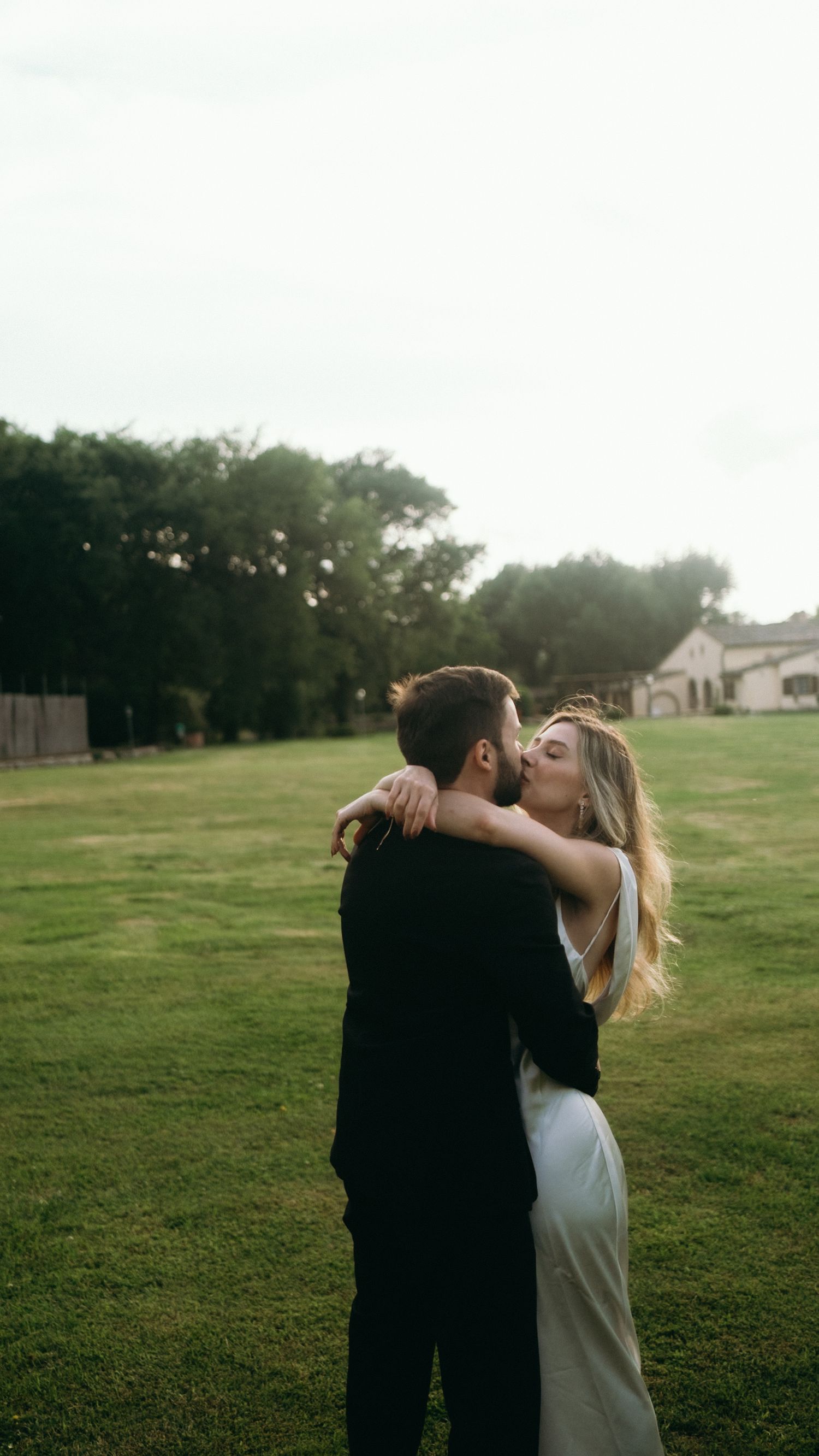 A couple in wedding attire sharing a romantic kiss on a lush green lawn with trees in the background.