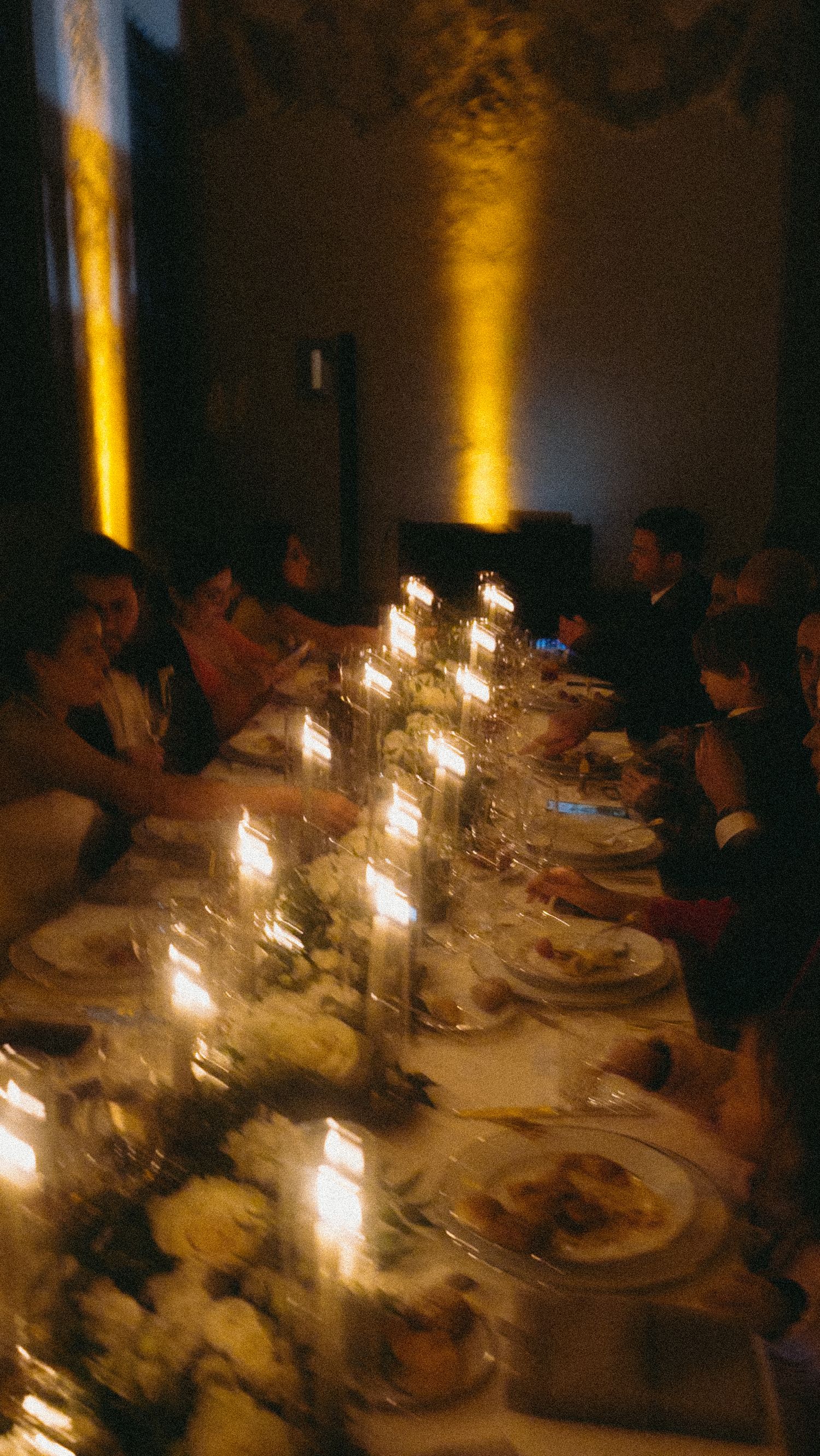 Guests dining by candlelight at long reception table with floral centerpieces in ornate venue.