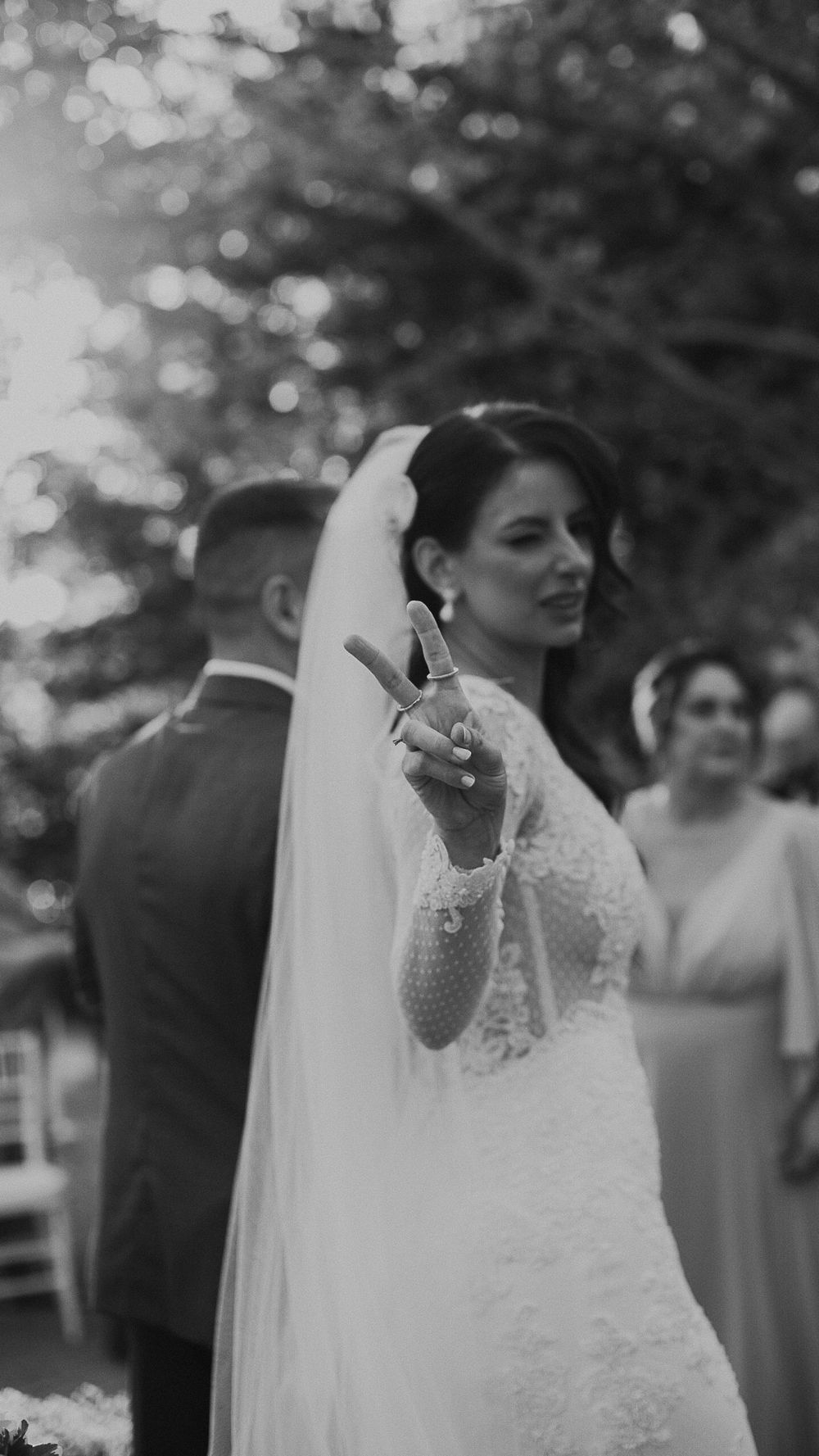 Bride in lace wedding gown and veil holds up wedding ring toward camera during outdoor ceremony in black and white.