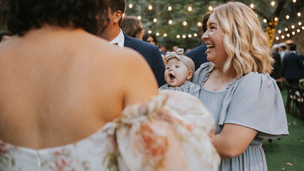 Wedding guest smiles while holding a delighted baby who reacts to meeting the bride outdoors.