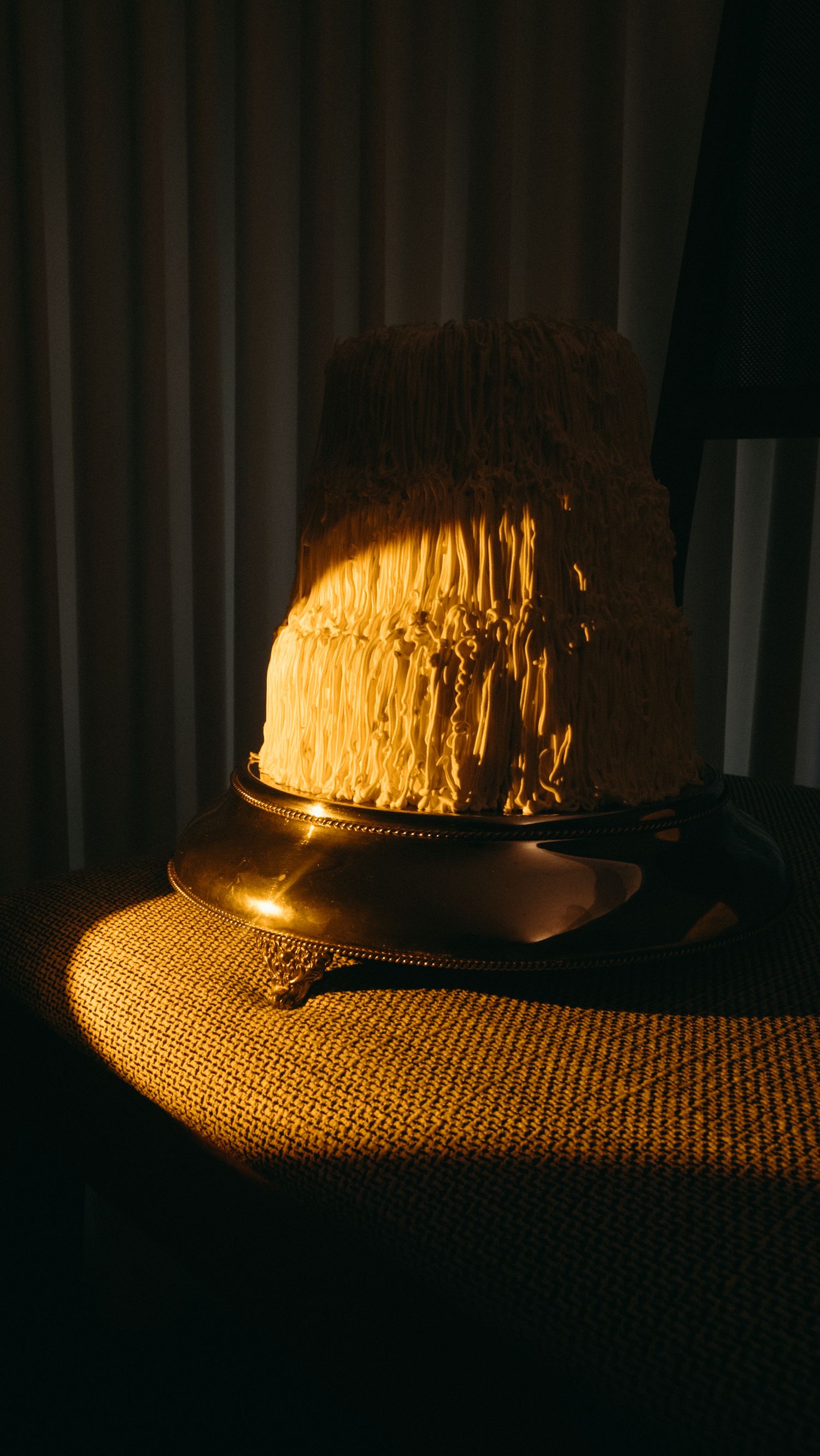 Two-tier white textured wedding cake dramatically lit by warm golden sunlight on a silver stand.