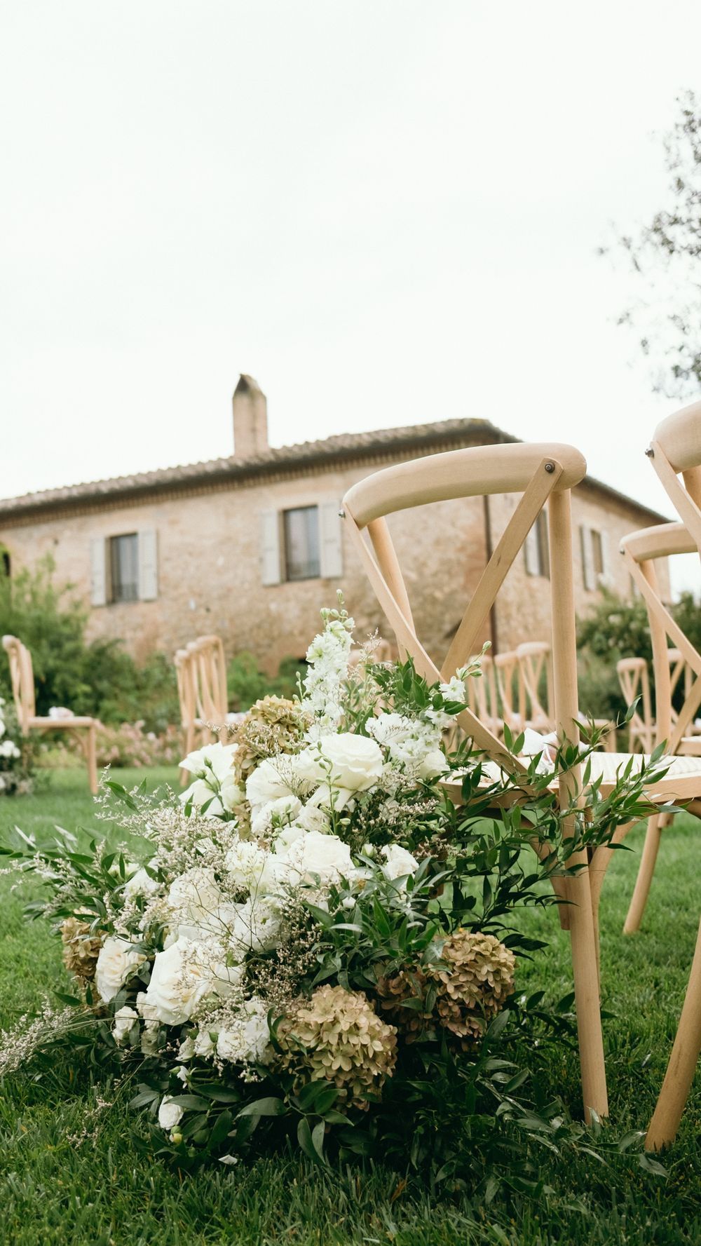 White floral arrangement with roses and hydrangeas decorating wooden chairs at an outdoor wedding ceremony.