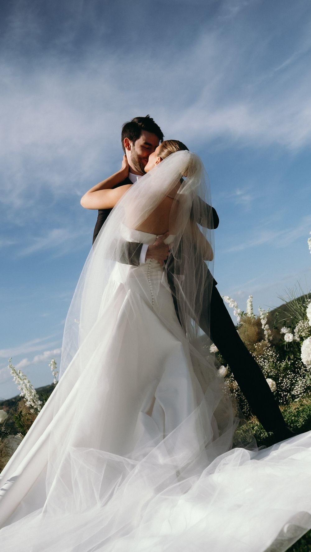 Groom lifting bride in flowing white gown and veil against blue sky at outdoor wedding ceremony.