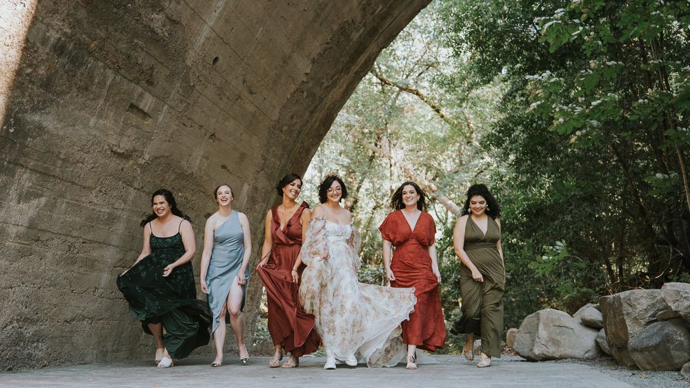 Six bridesmaids in colorful mismatched dresses walk under a stone arch bridge surrounded by lush greenery.