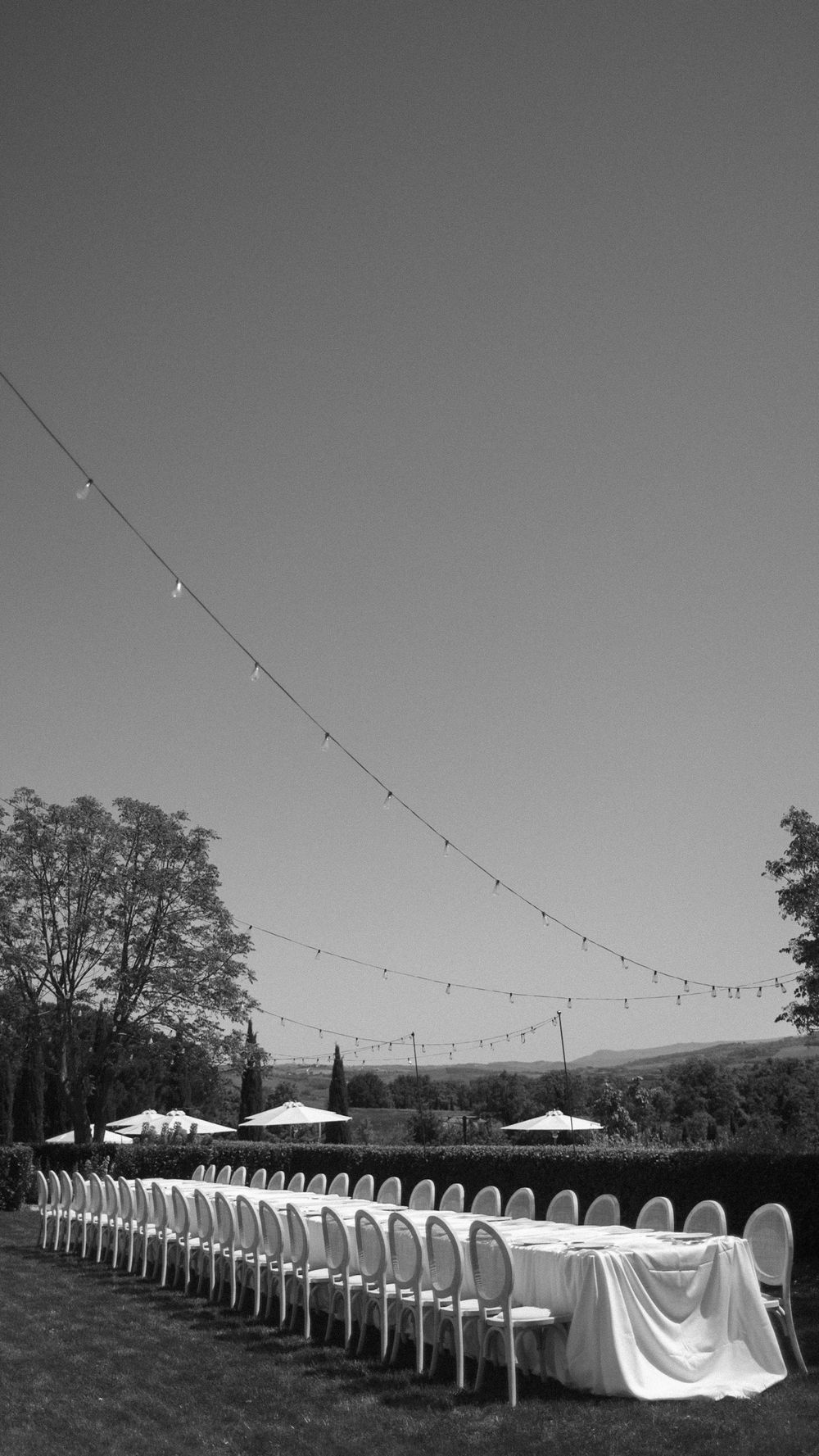 Black and white photo of long outdoor wedding reception table with chairs under string lights.