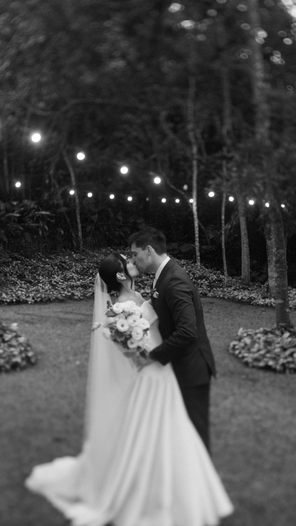 Black and white photo of bride and groom sharing first kiss under string lights in garden.
