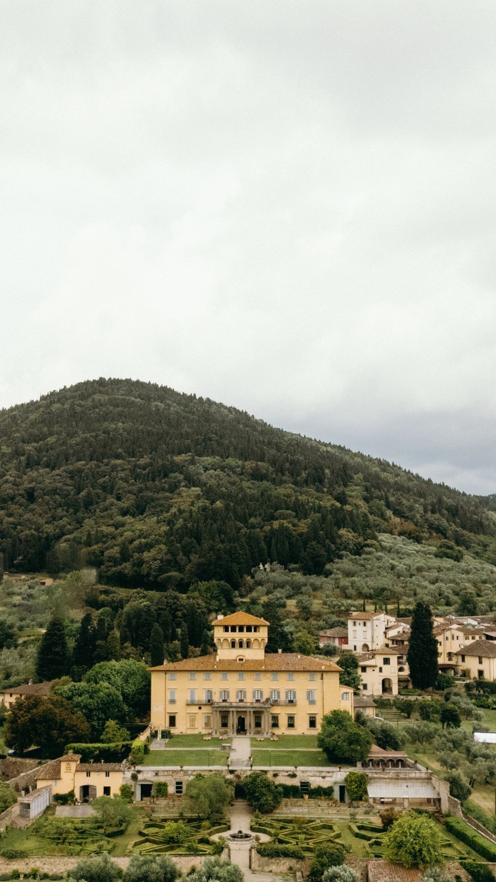 Aerial view of elegant Italian villa with formal gardens set against a lush green hillside under cloudy skies.