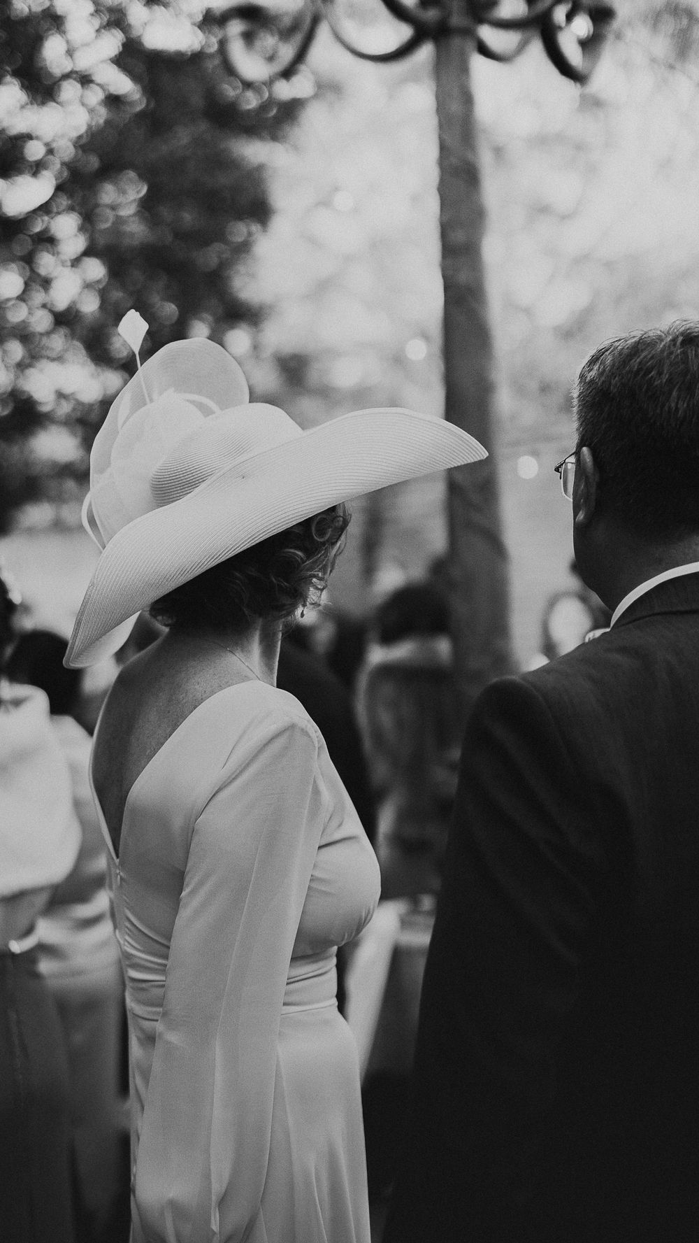 Elegant wedding guest in white wide-brim hat and backless gown attends outdoor garden wedding ceremony.