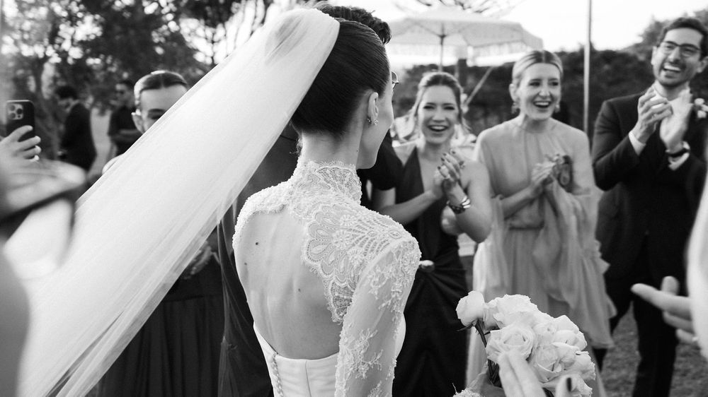 Bride in lace long-sleeve wedding gown with veil walks toward cheering wedding guests in black and white photo.