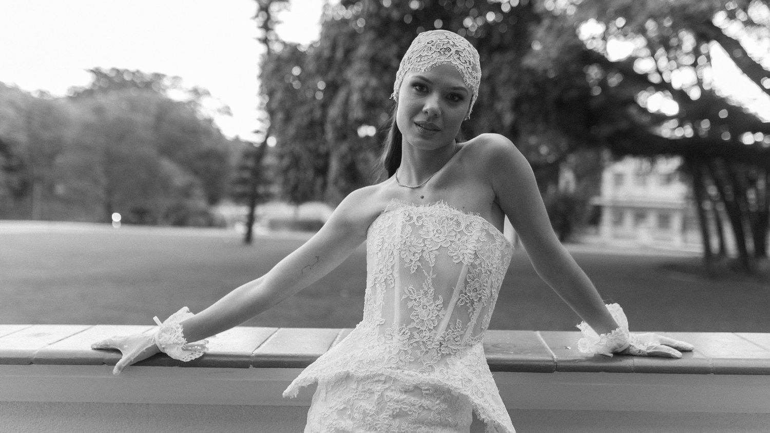 Black and white photo of an elegant woman in a lace wedding dress and headpiece posing outdoors.