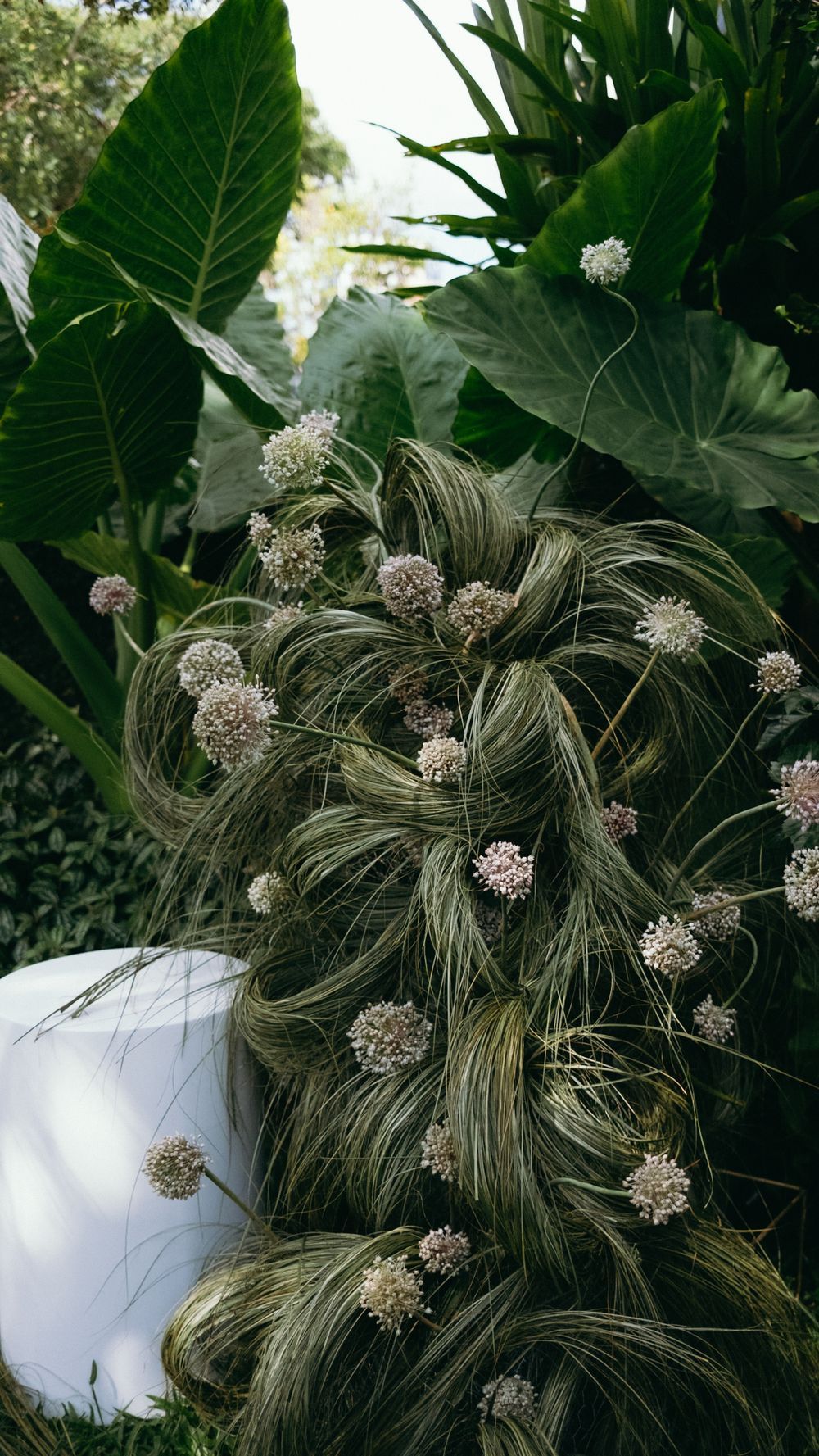 Bride's intricate updo hairstyle adorned with small white wildflowers and dried grasses against green foliage.