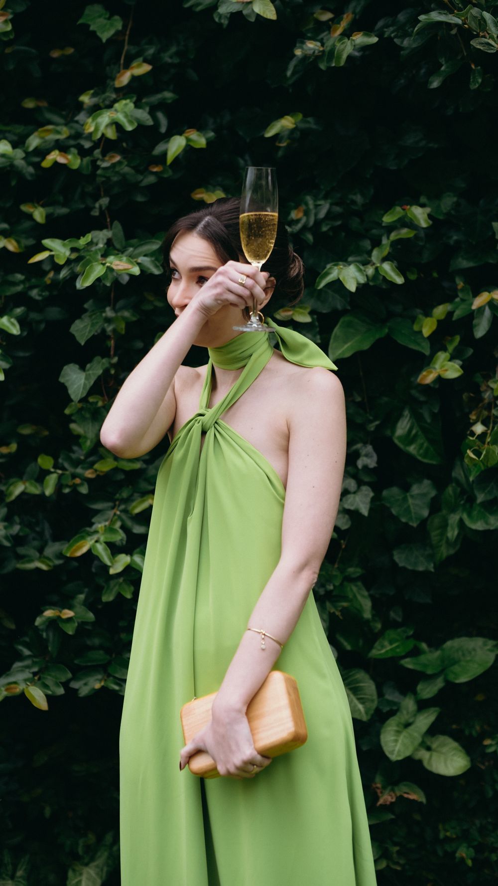 Woman in elegant green halter maxi dress holding champagne glass and clutch outdoors.
