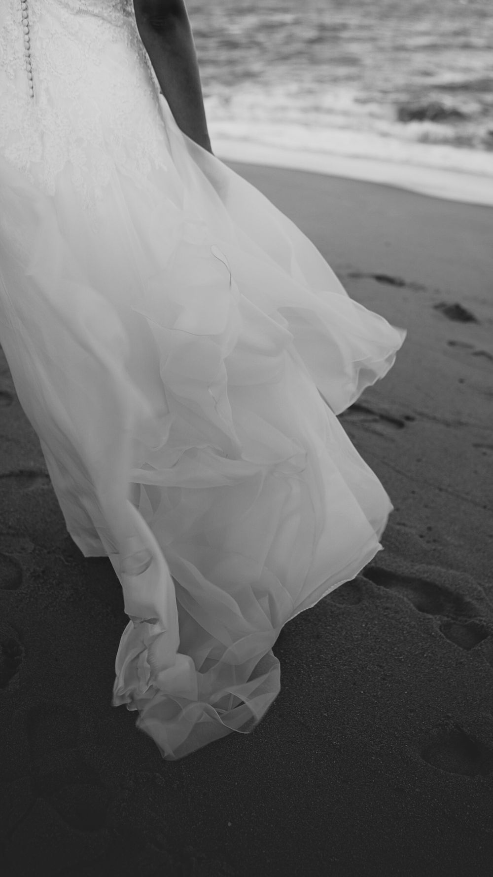 Black and white close-up of a wedding dress train flowing gracefully on a sandy beach near the ocean.