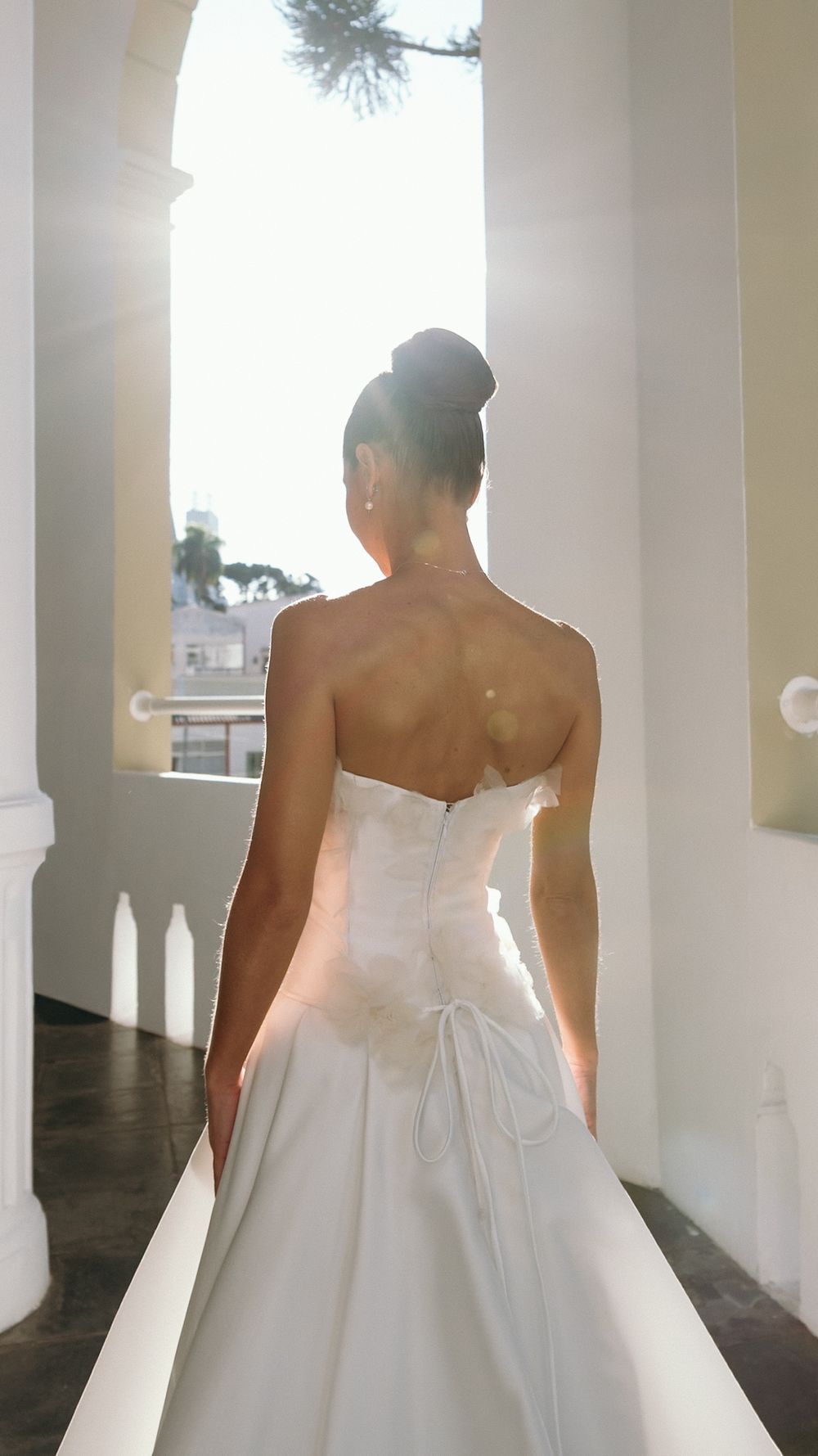 Bride in strapless white ball gown standing in sunlit archway with elegant updo hairstyle viewed from behind.