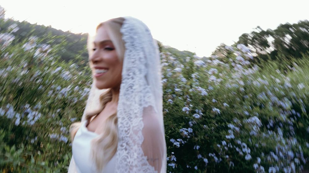 Smiling bride in a white gown and lace veil twirling among lush green flowering bushes outdoors.