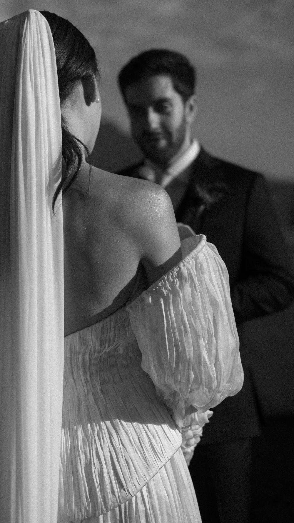 Black and white close-up of bride in off-shoulder pleated gown with veil facing groom during outdoor ceremony.