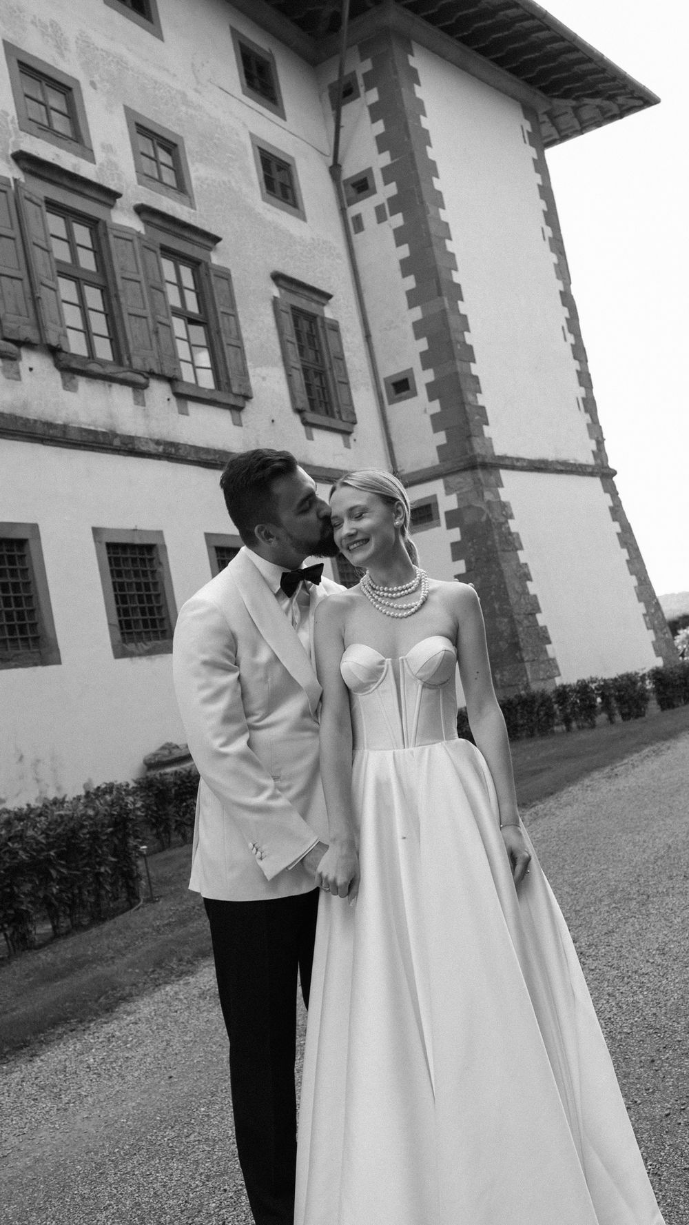 Black and white photo of groom kissing bride's cheek outside historic Italian villa.