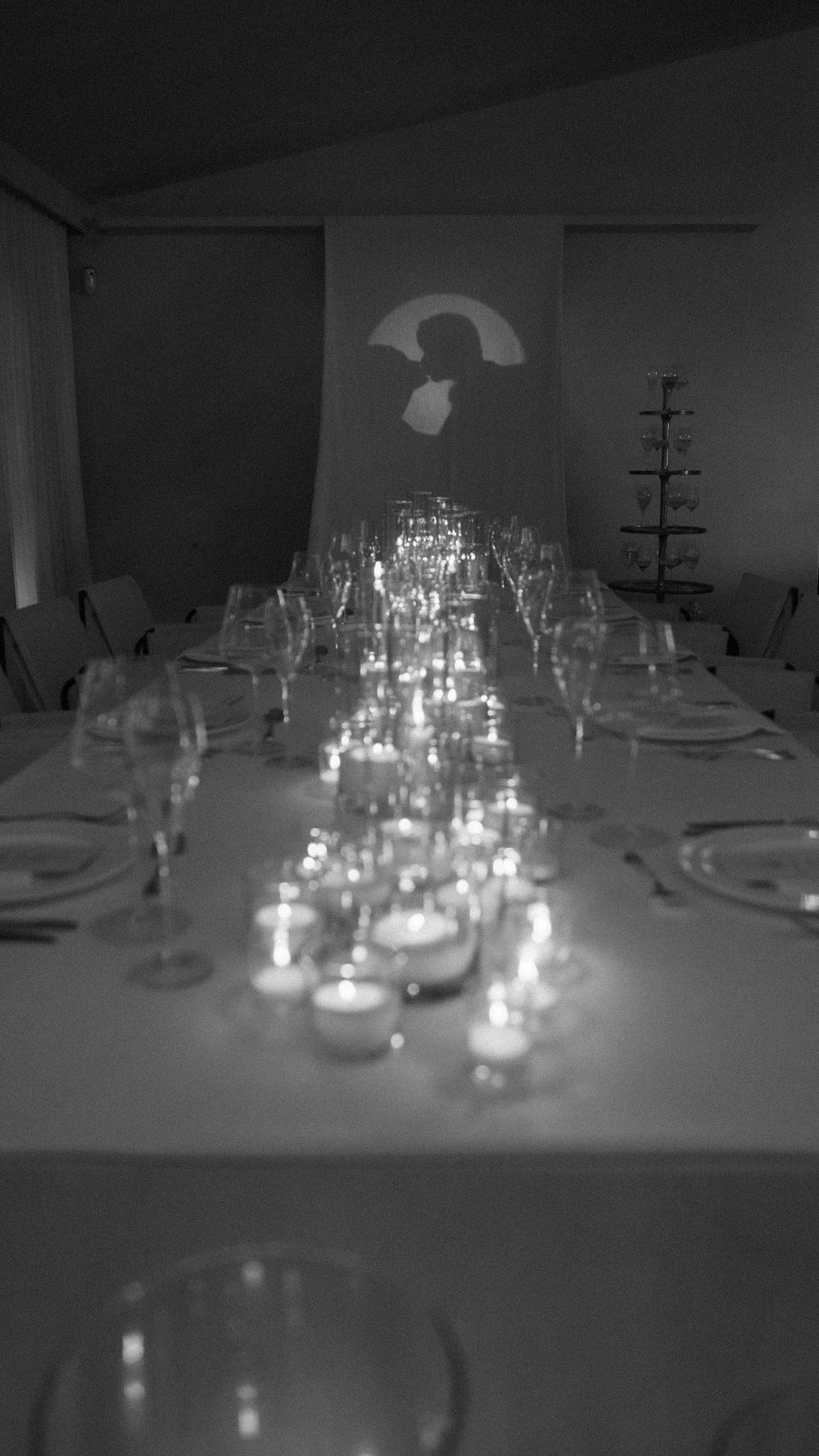 Black and white photo of candlelit wedding reception table with glassware and silhouette projection on wall.