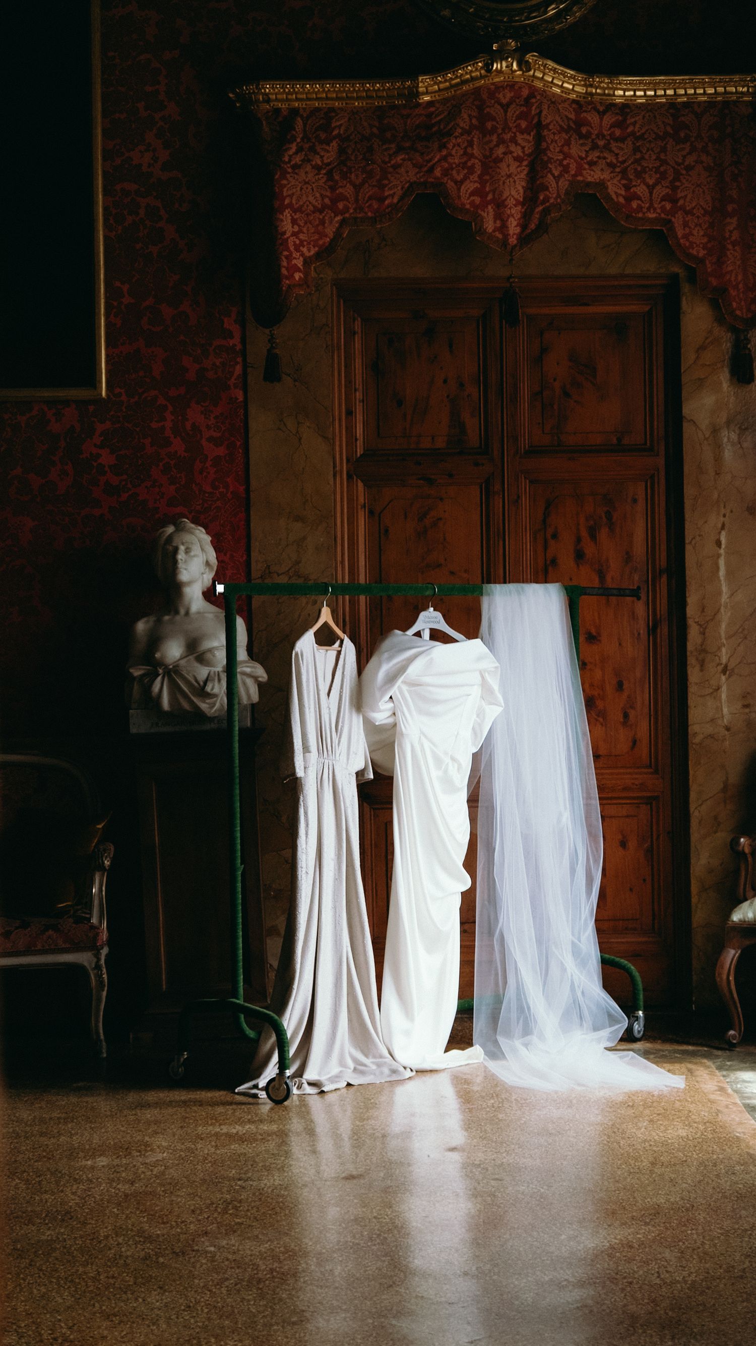 White wedding gown and veil hanging on a rack in an ornate vintage Venetian palace interior.