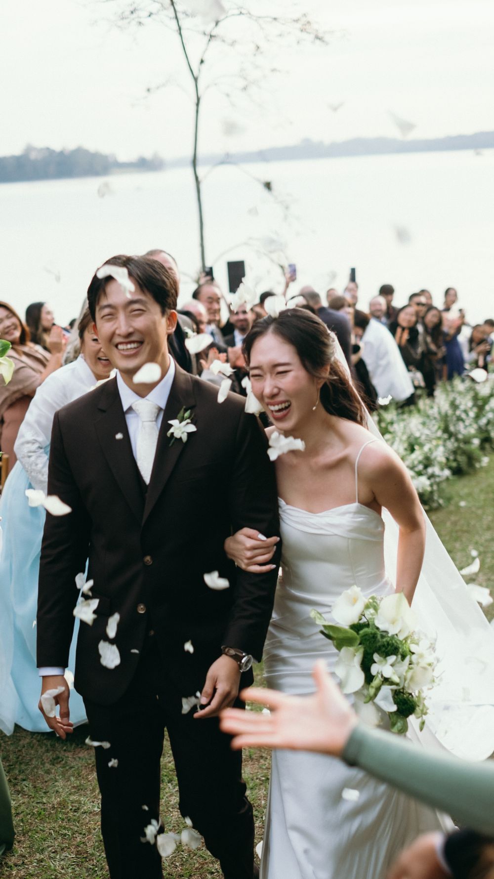 Joyful newlywed couple walks down aisle as guests shower them with white flower petals at outdoor lakeside wedding.
