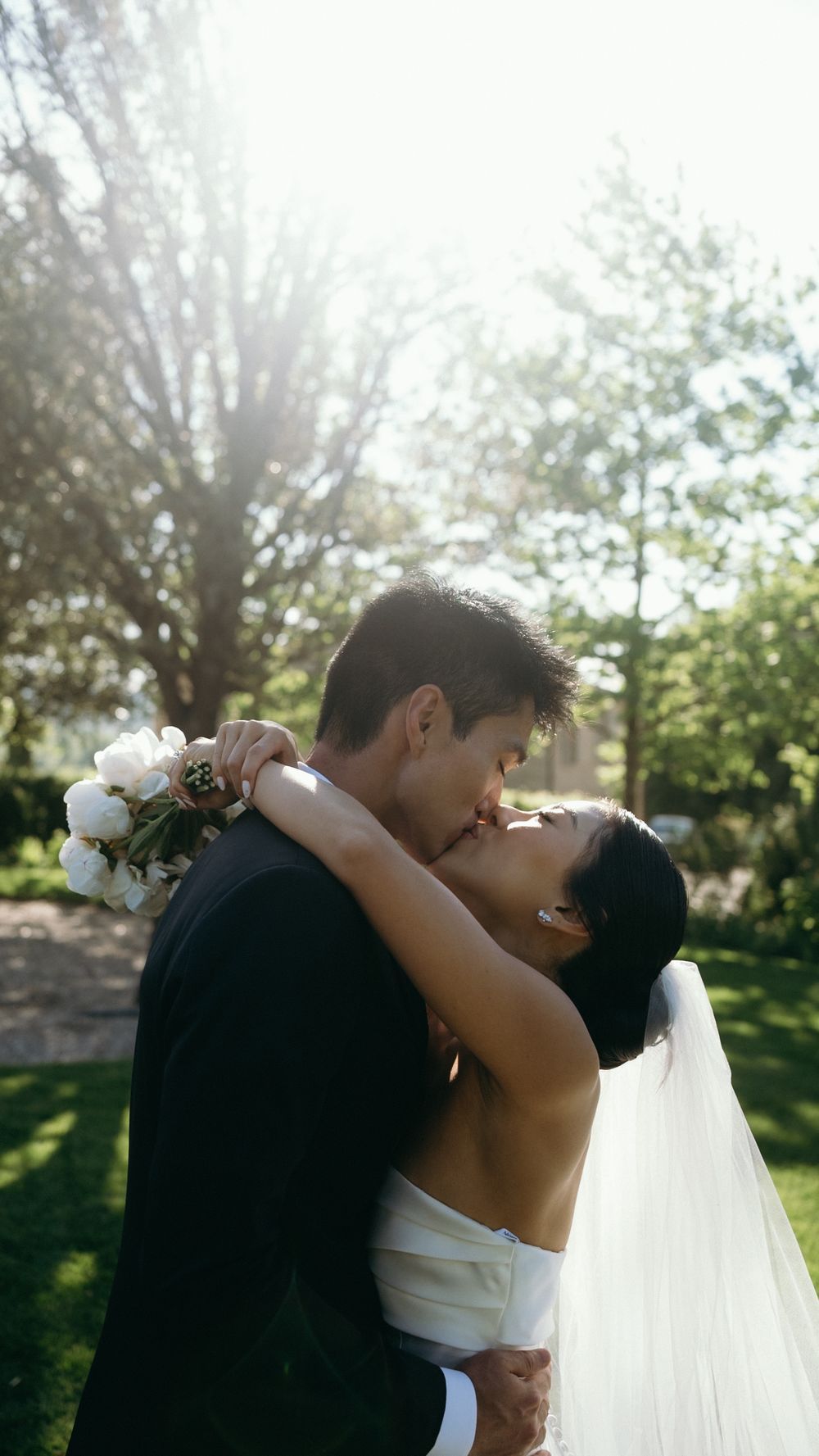 Bride and groom sharing passionate kiss outdoors with sunlight filtering through trees behind them.