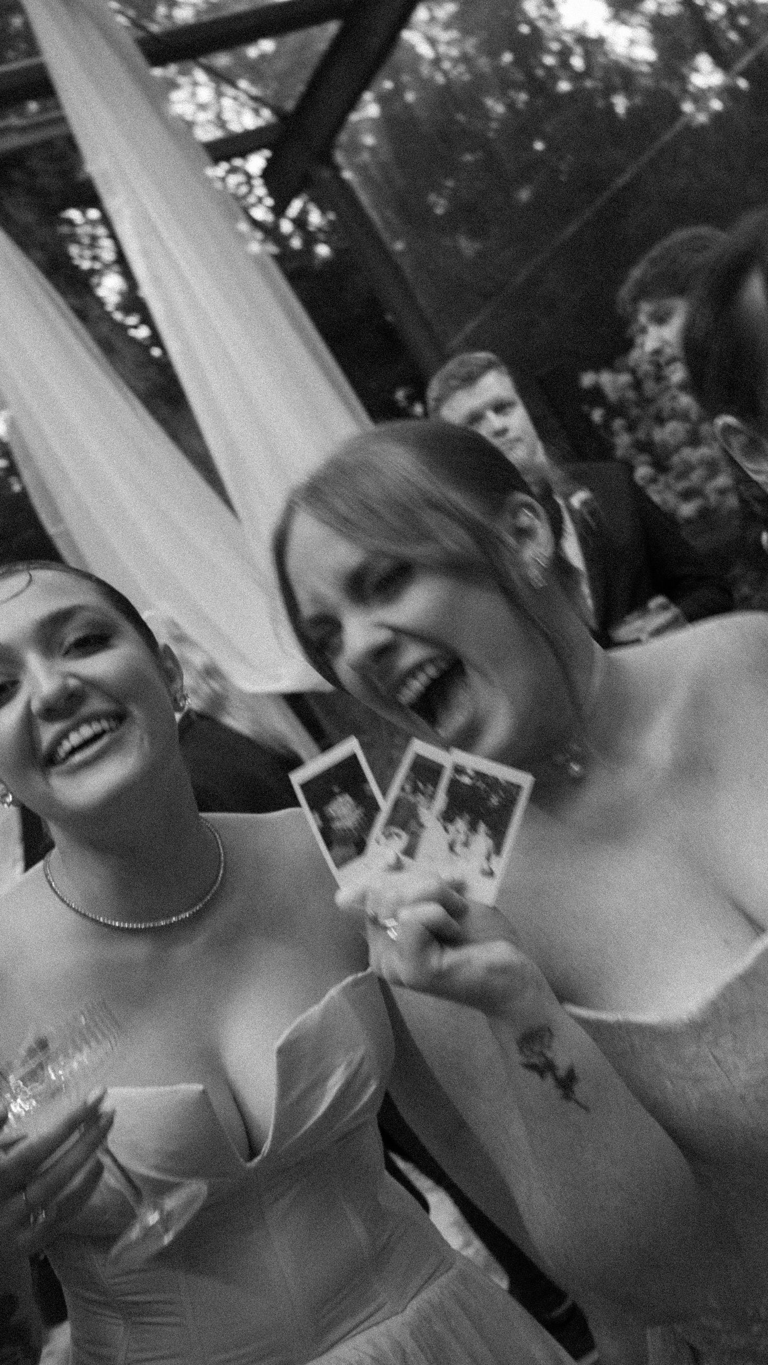 Black and white photo of two laughing women holding Polaroid photos at a wedding.