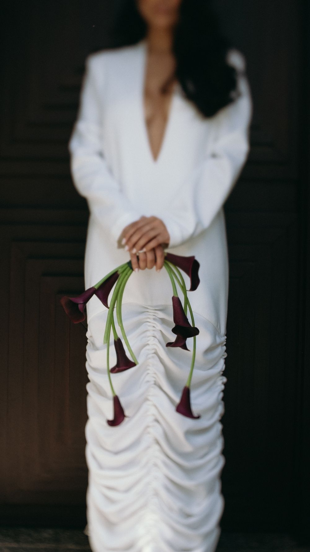 Bride in white ruched gown holding dark burgundy calla lily bouquet with long stems in dim interior.
