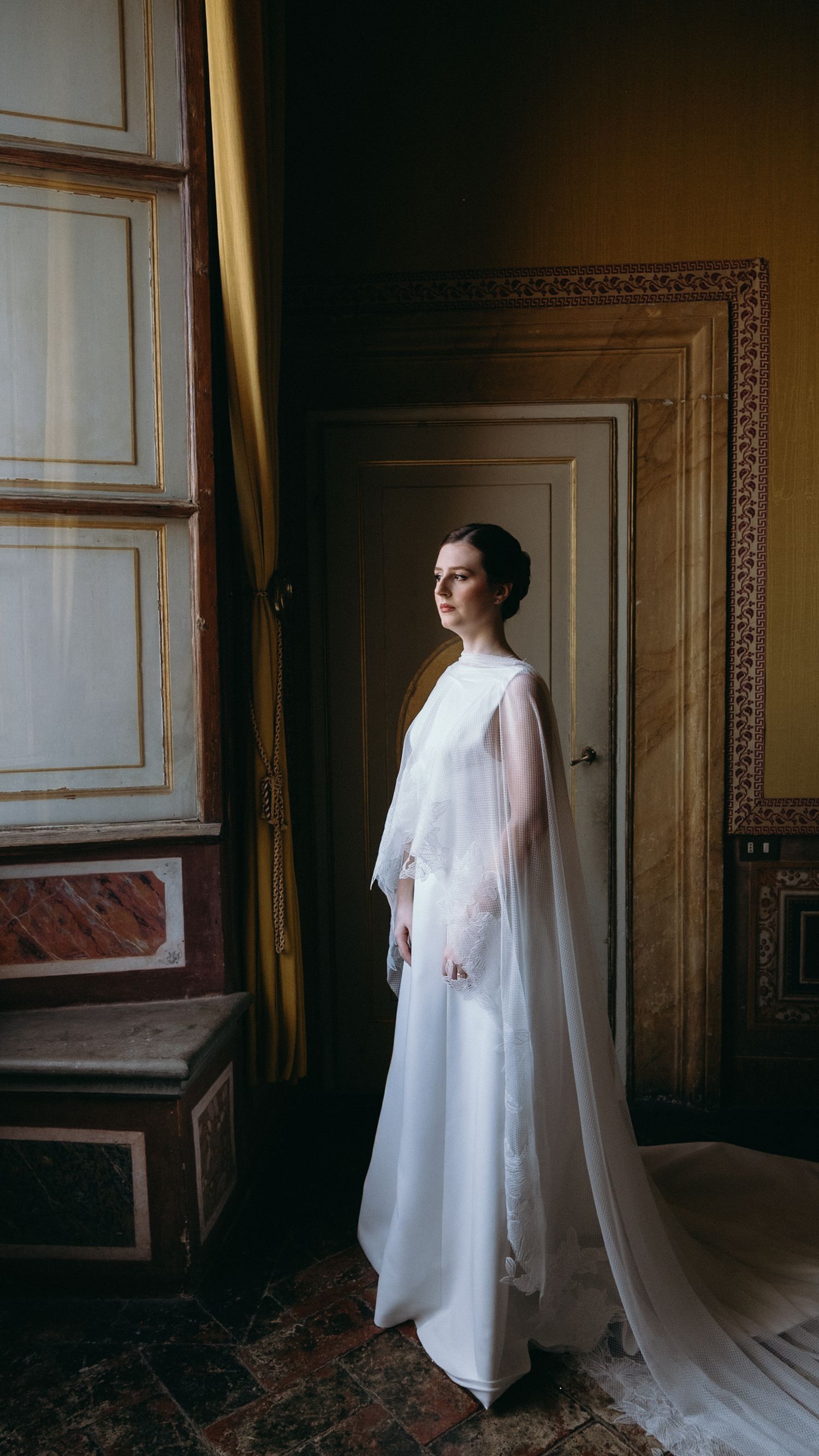 Elegant bride in minimalist white gown with sheer cape standing in ornate classical European palace interior.
