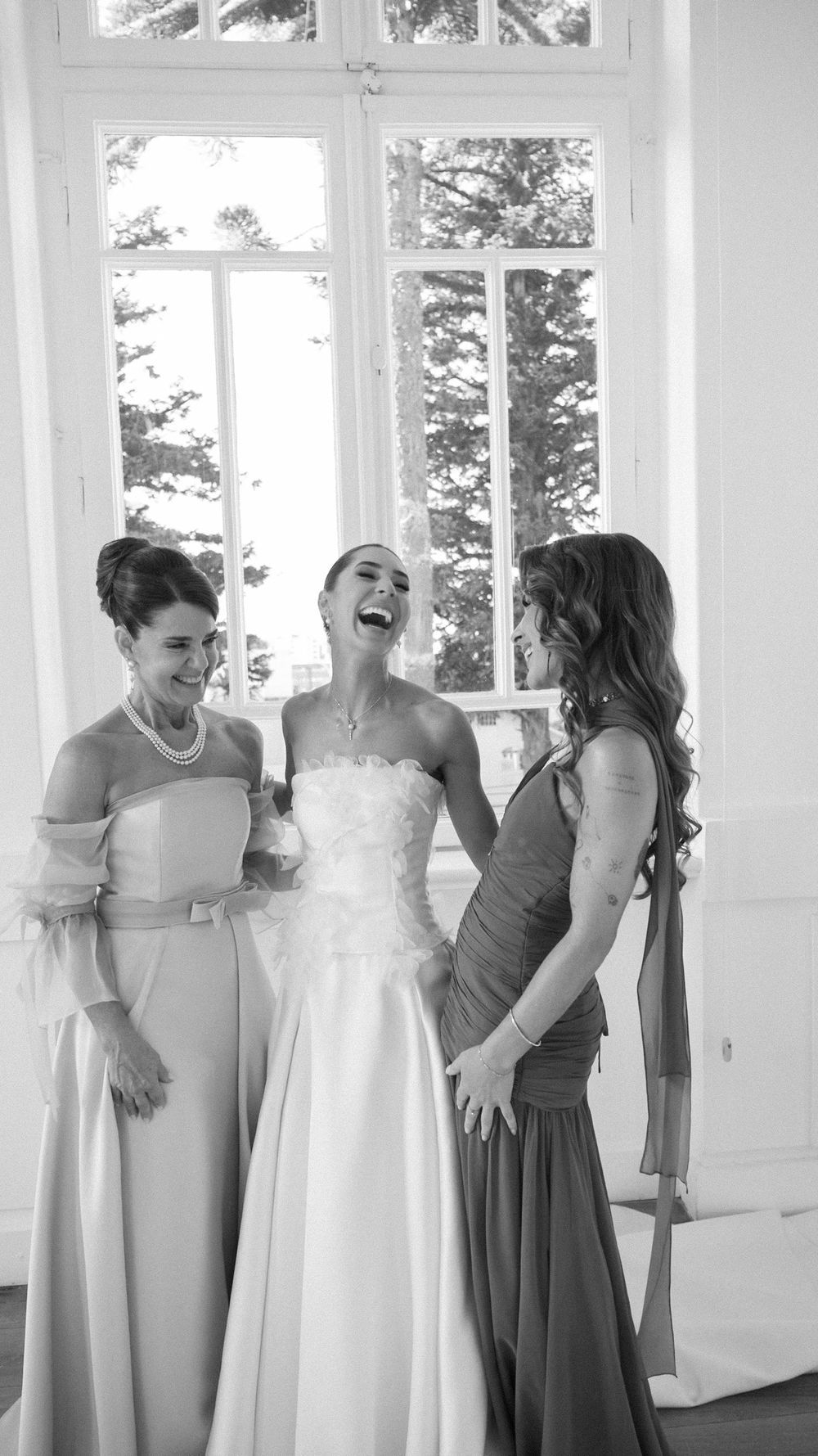 Bride laughing joyfully with two bridesmaids in elegant gowns inside a bright room with large windows.