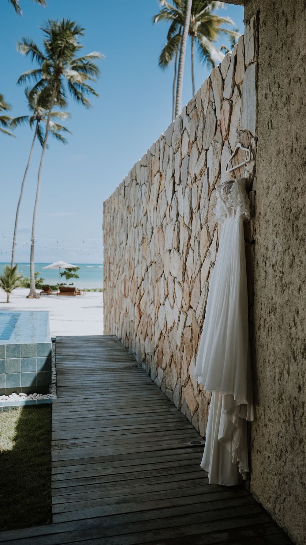 A white wedding dress hangs on a stone wall at a tropical beach resort with palm trees and ocean views.