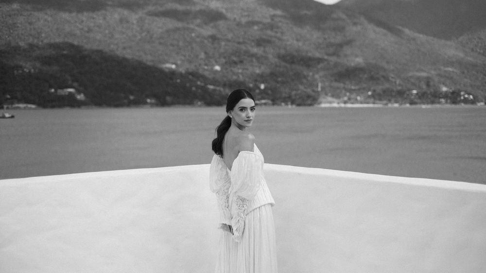 Bride in white off-shoulder gown posing gracefully on a terrace with mountain lake views in black and white.