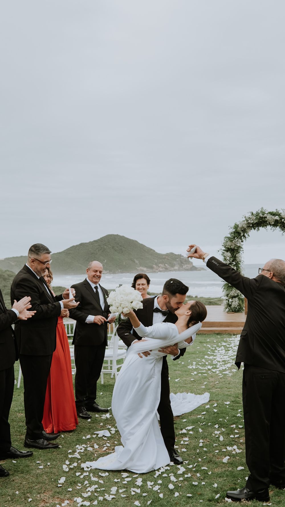 Groom dips bride for kiss at outdoor oceanside wedding ceremony with guests cheering.