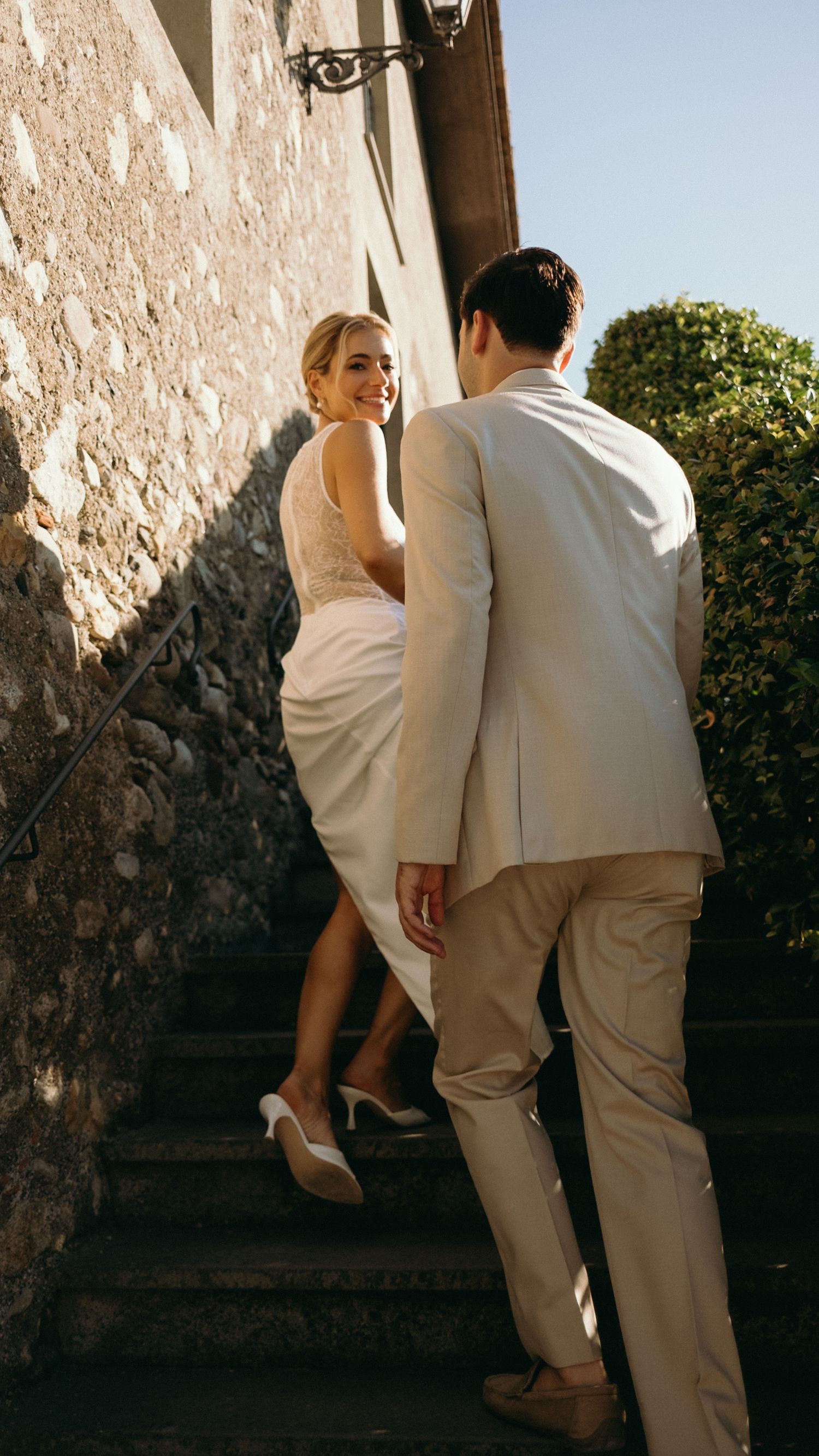 Smiling bride in white dress looks back at groom while climbing sun-drenched stone stairs.