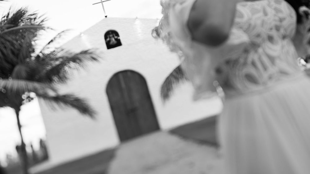 Black and white image of a bride approaching a white chapel with a bell tower and palm trees.