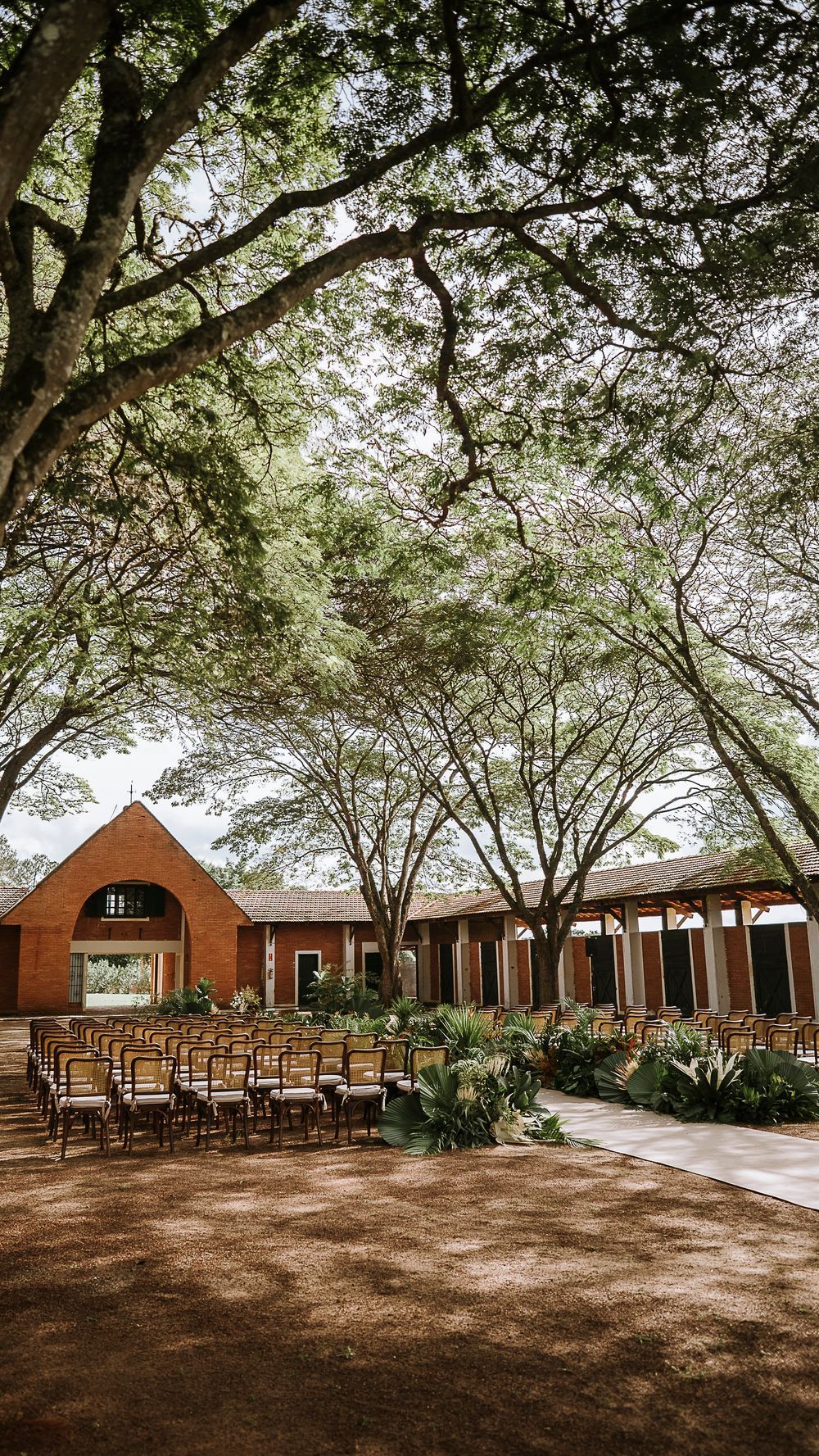 Elegant outdoor wedding ceremony setup with wooden chairs arranged under large trees at rustic brick venue.