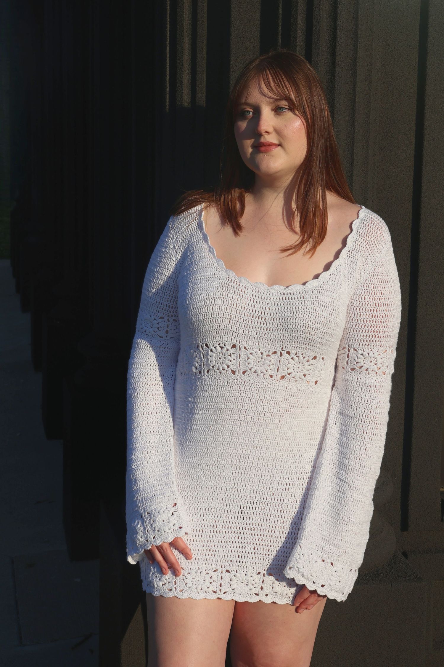 A woman in a white knit long-sleeve dress posing outdoors with direct sunlight illuminating half of her face and dress.