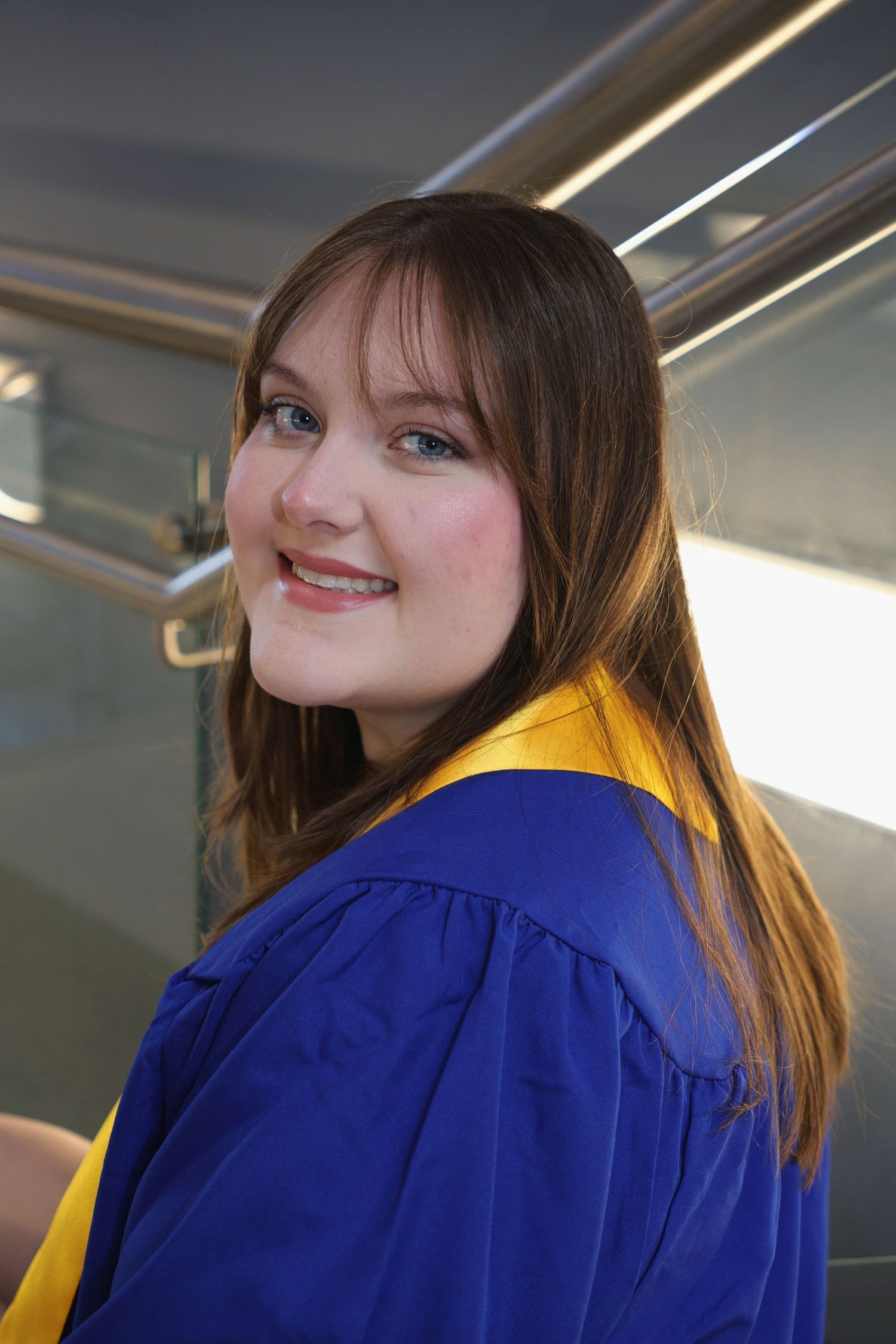 A close-up portrait of a smiling woman with bangs wearing a blue graduation gown and a yellow stole indoors.
